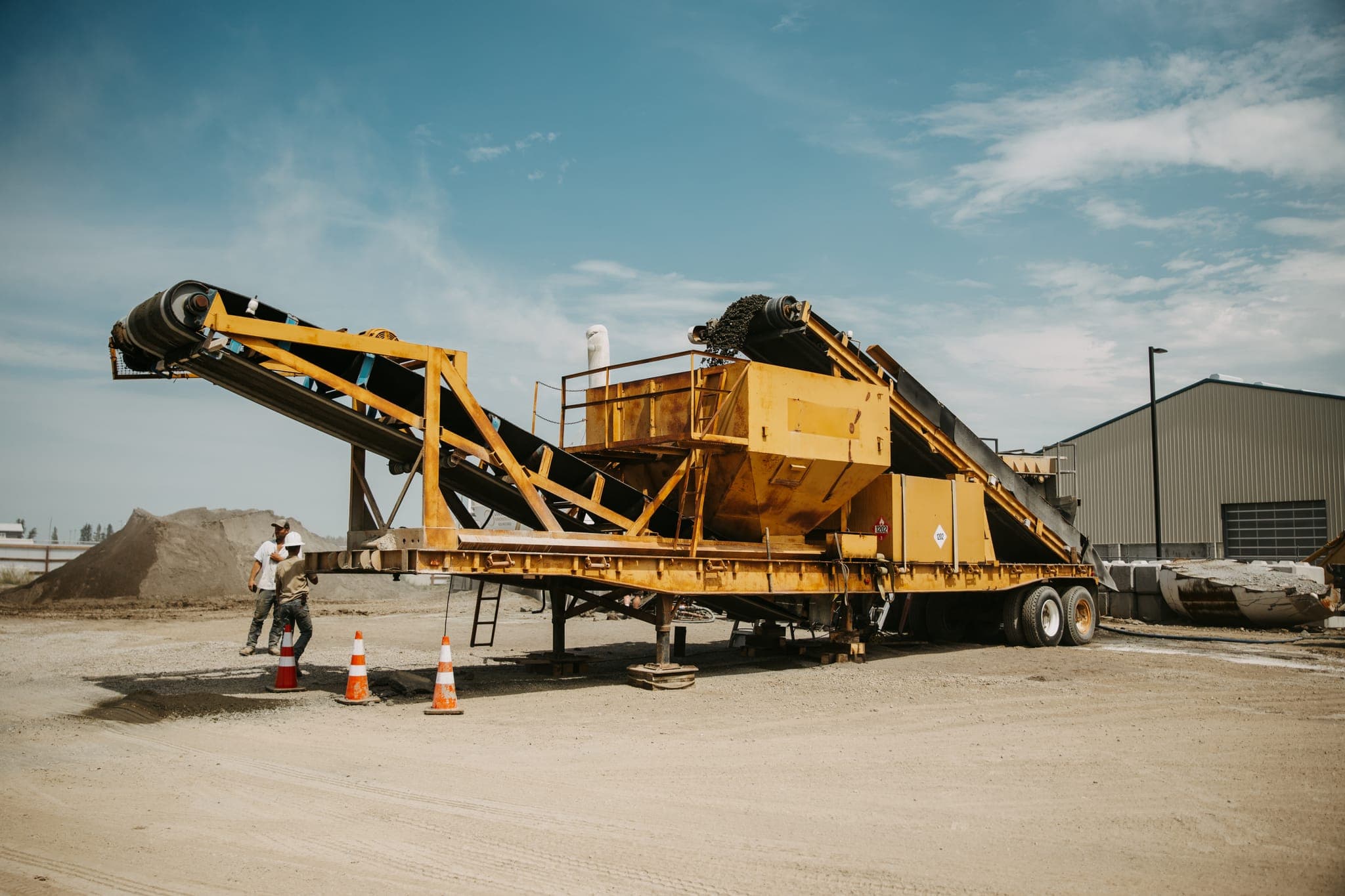 Two crew members in hard hats beside the pugmill trailer setup, safety cones marking the area.