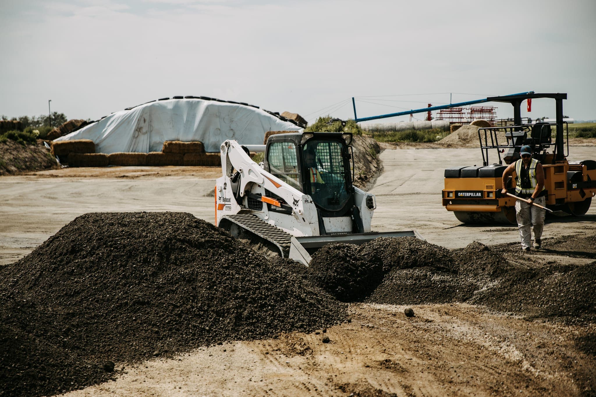 Skid steer spreading roller-compacted concrete material while a vibratory roller compacts nearby, crew member raking, feedlot site.