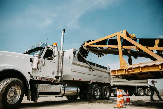 Wide yard view of a dump truck being loaded with RCC mix, a worker and second truck nearby.