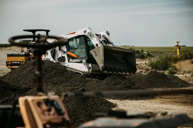 Skid steer bucket digging into a material pile, a Caterpillar roller and GPS tripod in the background.